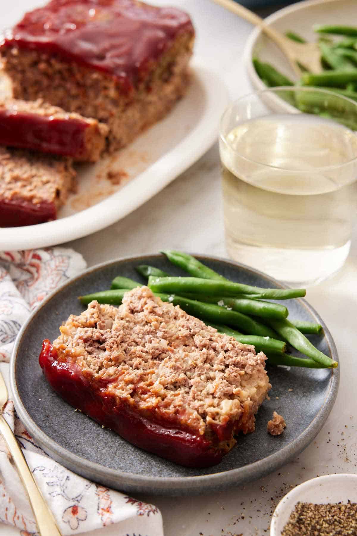 A slice of meatloaf with green beans on a plate with a glass of wine and the rest of the meatloaf in the background.