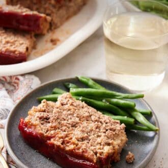 Pinterest graphic of a slice of meatloaf with green beans on a plate with a glass of wine and the rest of the meatloaf in the background.