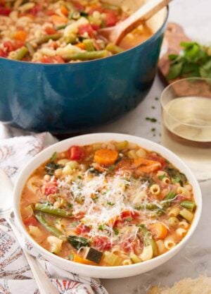 A bowl of minestrone soup with a glass of wine and pot of soup in the background.