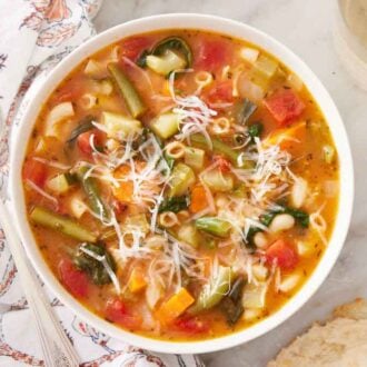 Overhead view of a bowl of minestrone soup topped with parmesan on top. Some torn bread and glass of wine off to the side.