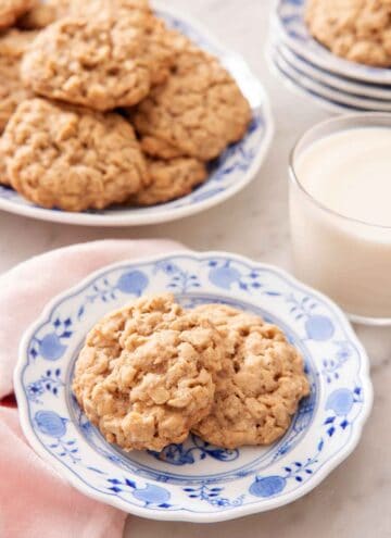 A plate with two oatmeal cookies with a glass of milk and additional cookies in the background.