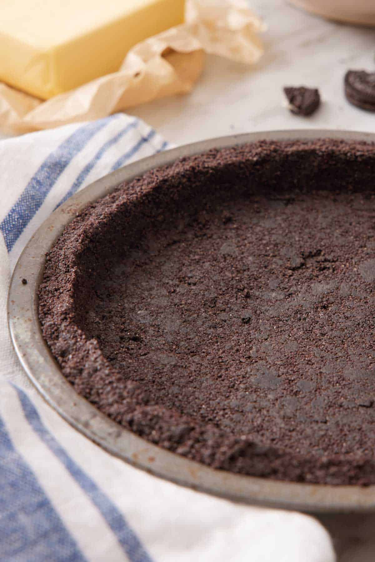 An Oreo pie crust in a pie pan with a stick of butter out of focus in the background.