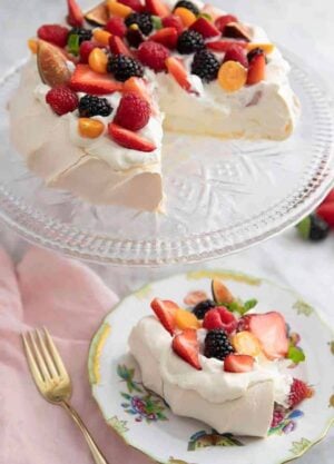 A plate with a slice of pavlova topped with berries with the rest of the pavlova on a clear cake stand.