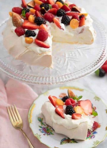A plate with a slice of pavlova topped with berries with the rest of the pavlova on a clear cake stand.