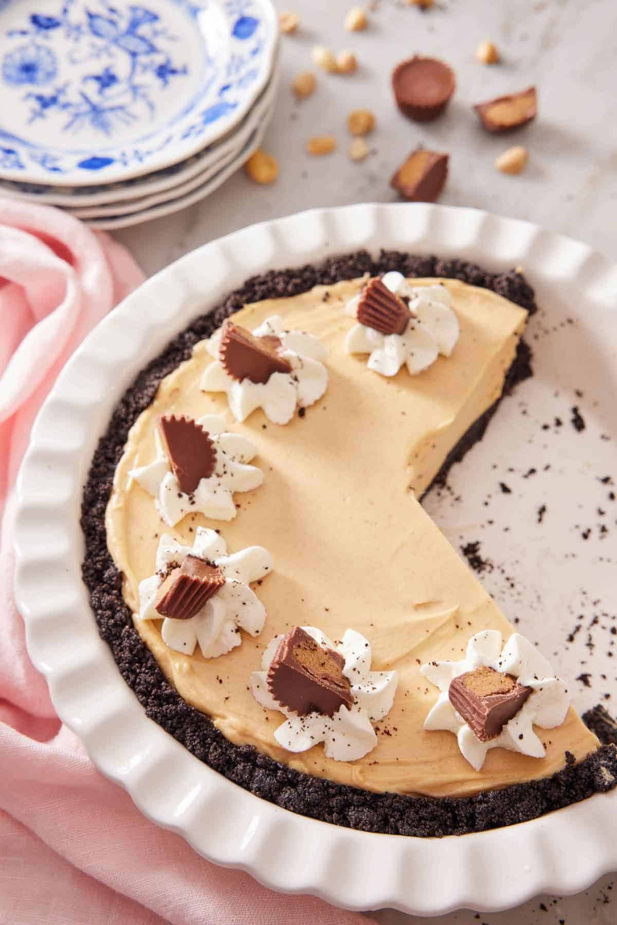 A peanut butter pie in a white baking dish with a couple of slices cut out. Whipped cream and Reese's garnished on to.