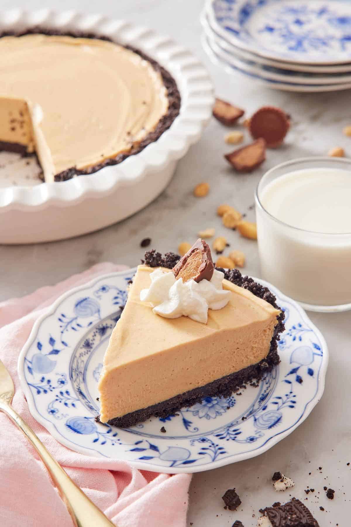 A slice of peanut butter pie with a glass of milk and the rest of the pie in the background.