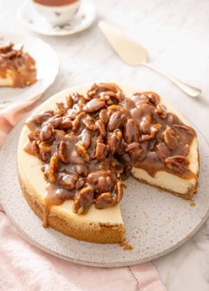 A pecan pie cheesecake with a slice taken out. A cake spatula and a cup of tea in the background.
