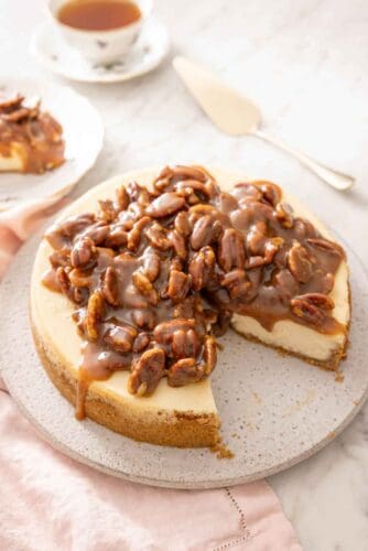 A pecan pie cheesecake with a slice taken out. A cake spatula and a cup of tea in the background.