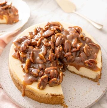 A pecan pie cheesecake with a slice taken out. A cake spatula and a cup of tea in the background.