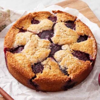 A plum cake on a parchment lined wooden serving board with two plums beside it.