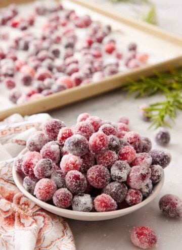 A bowl of sugared cranberries with a sheet pan with more in the background.