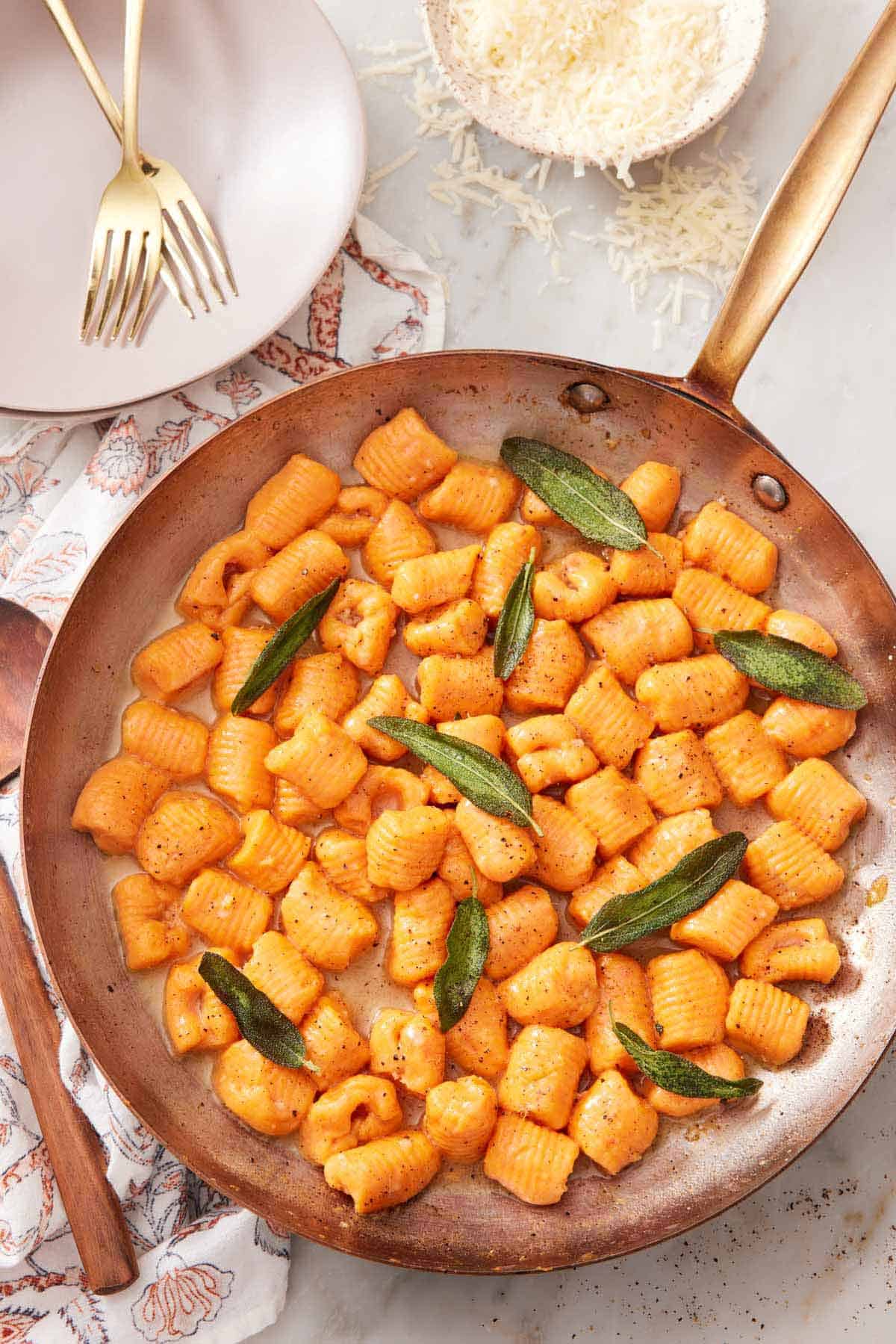 Overhead view of a skillet of sweet potato gnocchi with sage. A bowl of parmesan on the side along with plates and forks.
