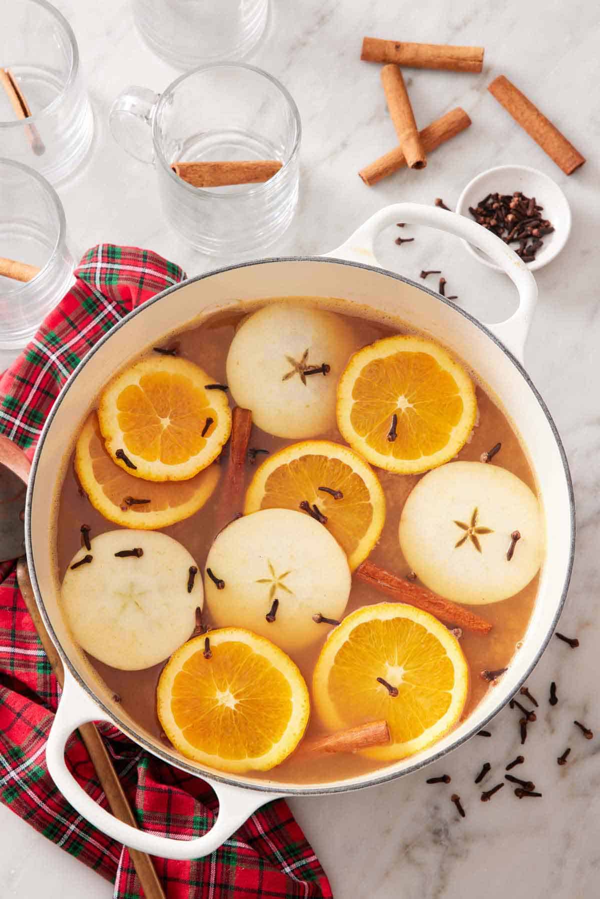 Overhead view of a white dutch oven of wassail topped with sliced orange, apples, cloves, and cinnamon sticks. Surrounded by empty glasses and more ingredients.