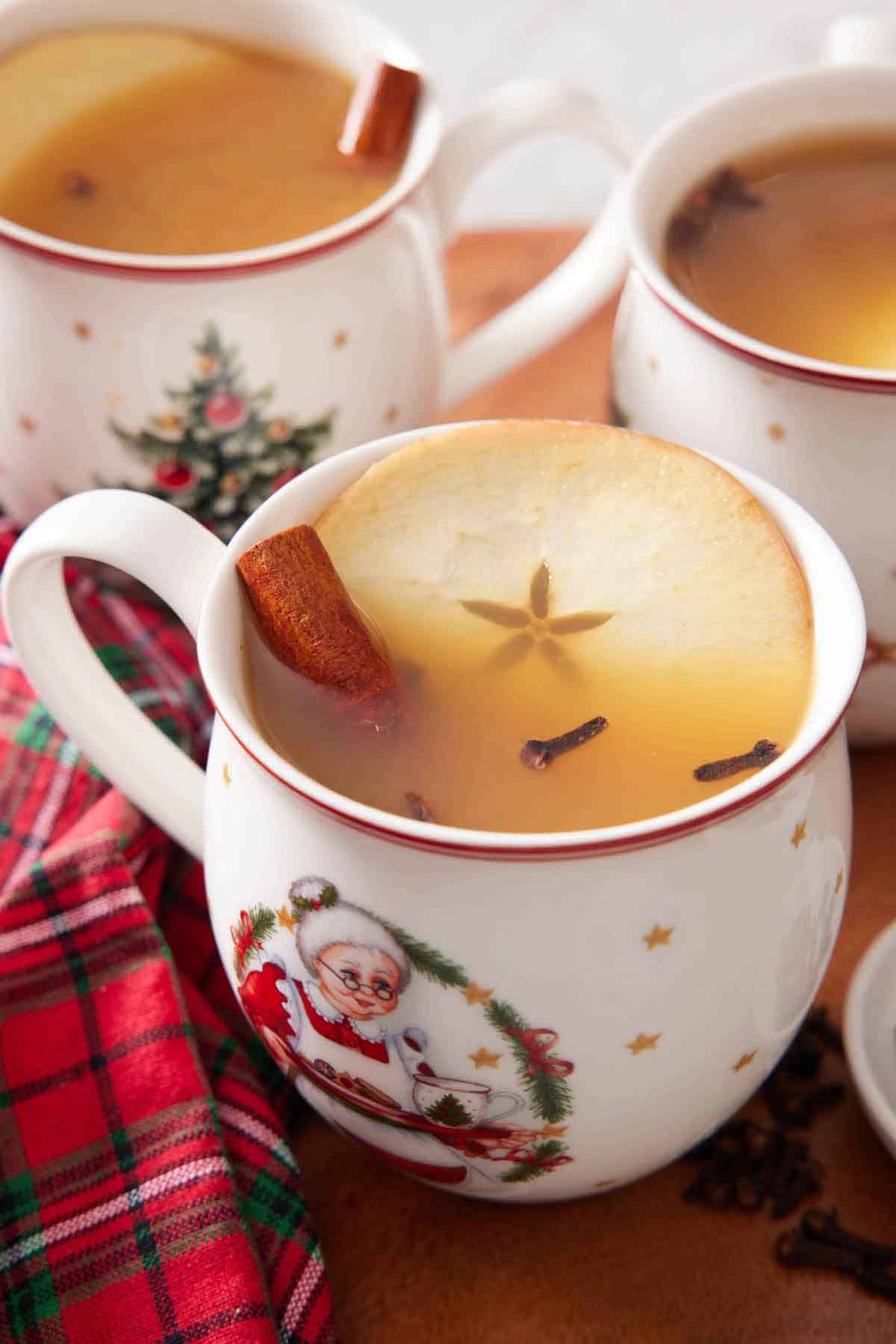 A festive mug of wassail with a cinnamon stick, apple slice, and cloves. More mugs in the back.