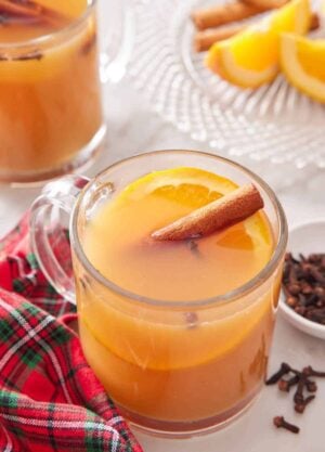 A glass of wassail with a cinnamon stick and slice of orange. A linen, cloves, cut oranges, and another glass of wassail off to the side.