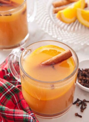 A glass of wassail with a cinnamon stick and slice of orange. A linen, cloves, cut oranges, and another glass of wassail off to the side.