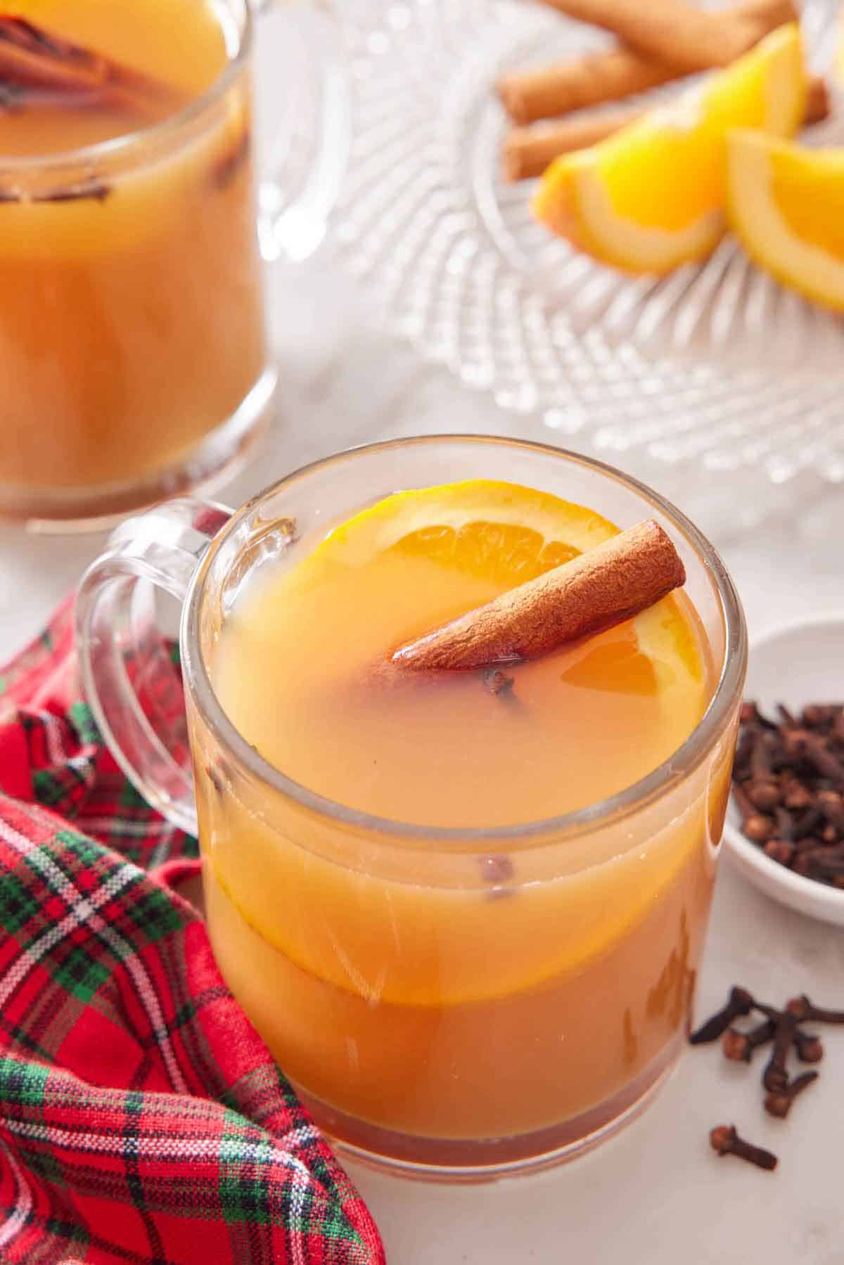 A glass of wassail with a cinnamon stick and slice of orange. A linen, cloves, cut oranges, and another glass of wassail off to the side.