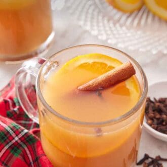 Pinterest graphic of a glass of wassail with a cinnamon stick and slice of orange surrounded by a linen napkin, cloves, cut oranges, and another mug.