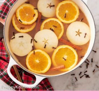 Pinterest graphic of an overhead view of a pot of wassail topped with sliced orange, apples, cloves, and cinnamon sticks.