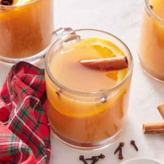 A glass of wassail with a cinnamon stick and orange slice. More glasses off to the side with a linen napkin, cinnamon sticks, and cloves beside it.