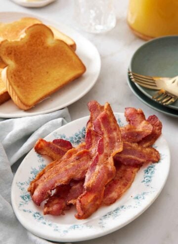 A platter of air fryer bacon with a plate of toast in the background along with a stack of plates and forks.