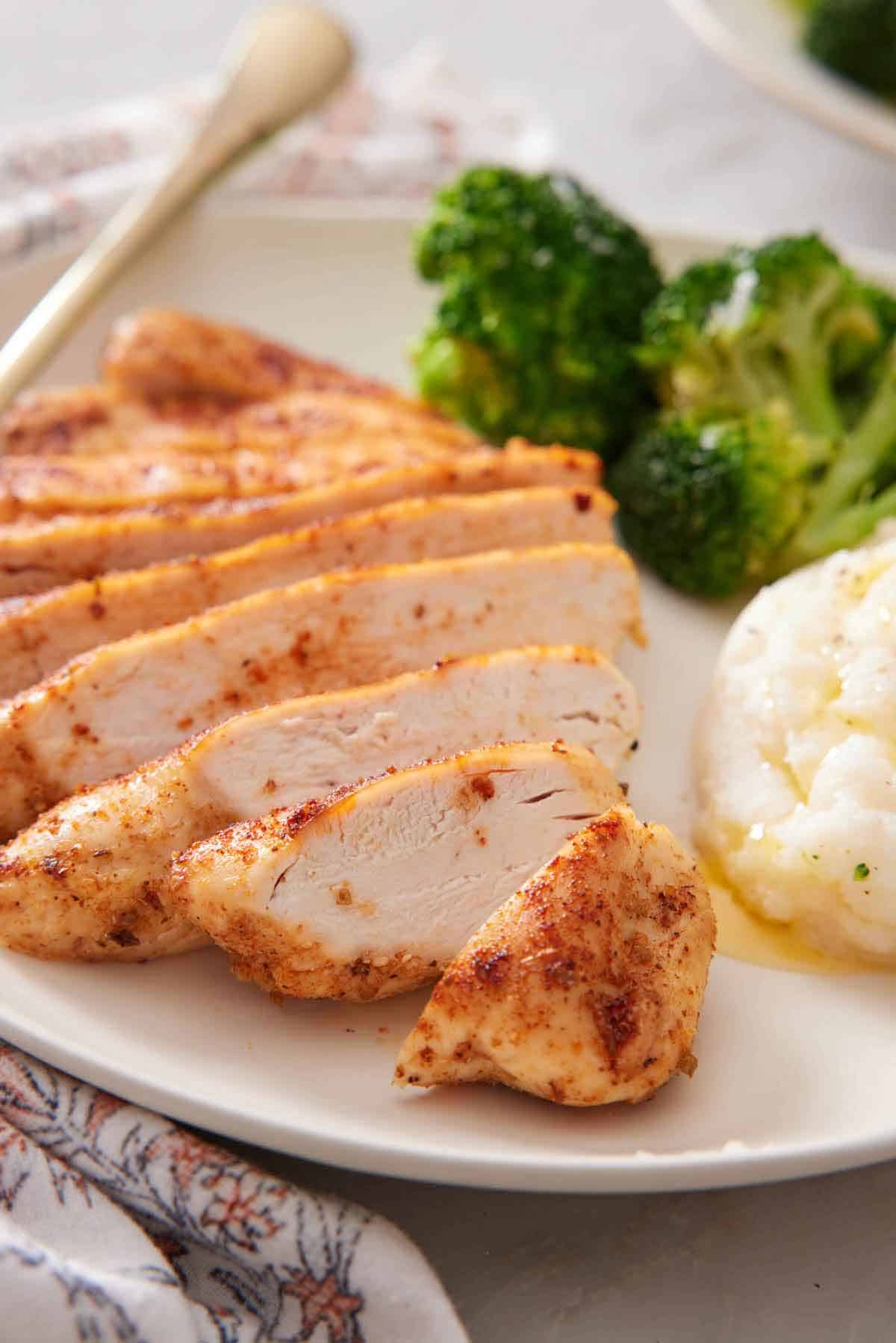 A close up of sliced air fryer chicken breast on a plate with some mashed potatoes and broccoli on the side.