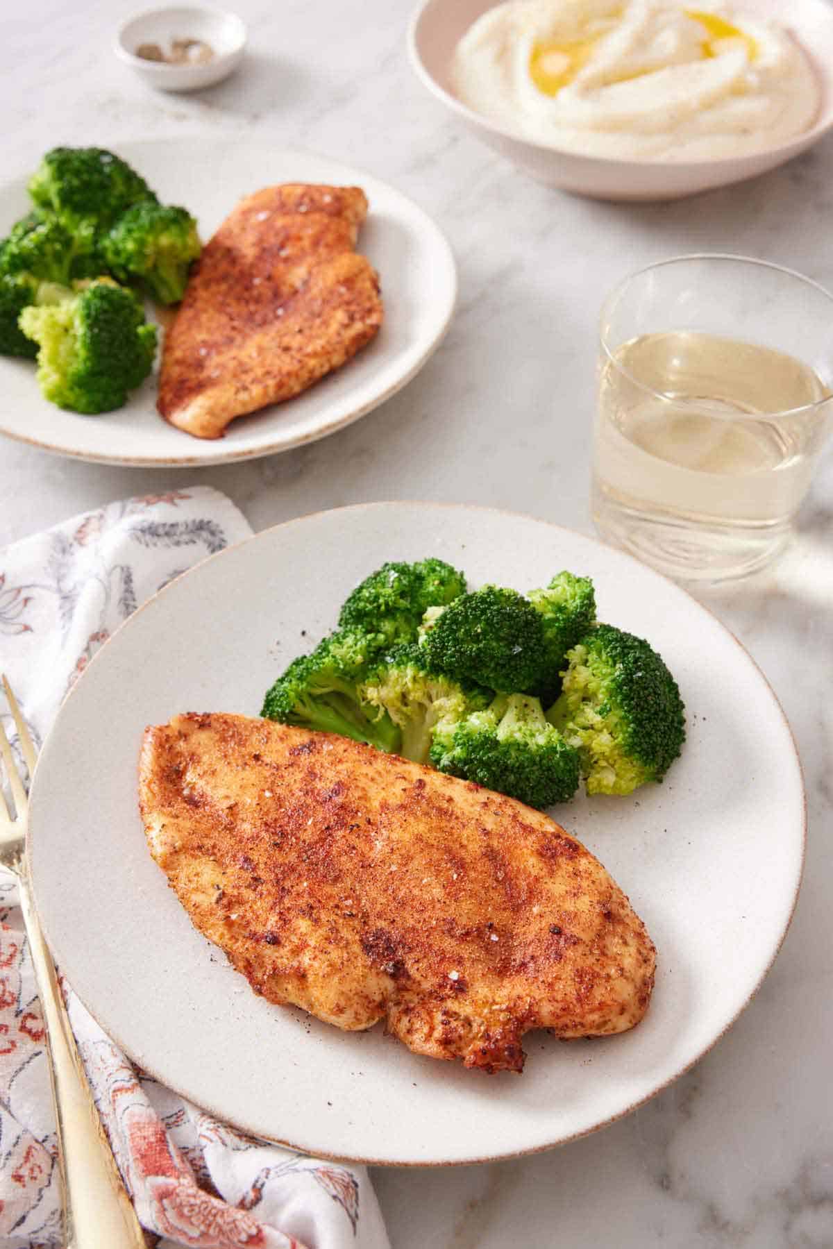 Two plates of air fryer chicken breasts with a side of broccoli. A drink and bowl of mashed potatoes in the background.