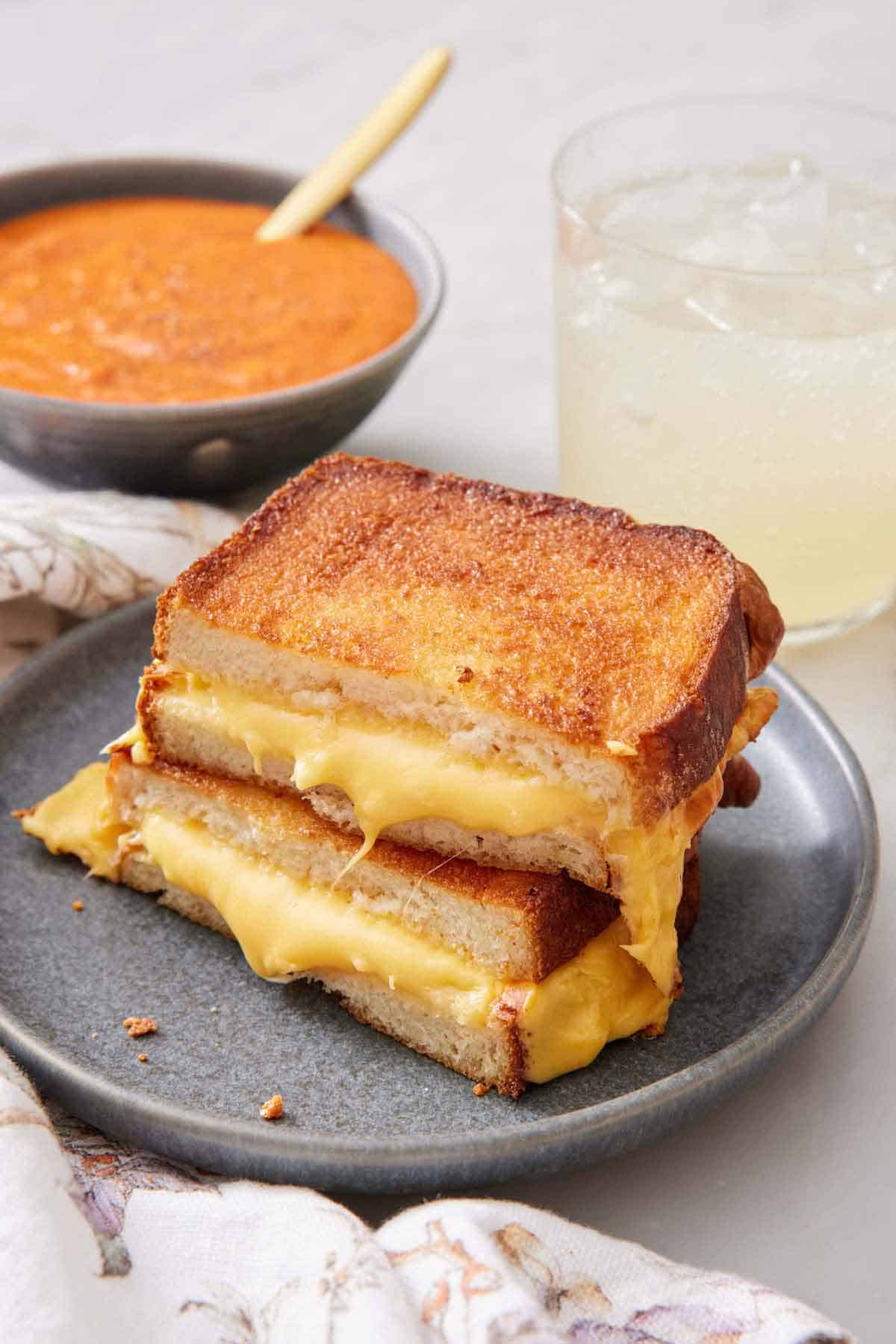 A plate with an air fryer grilled cheese cut in half, stacked on top of each other. A drink and bowl of tomato soup in the back.