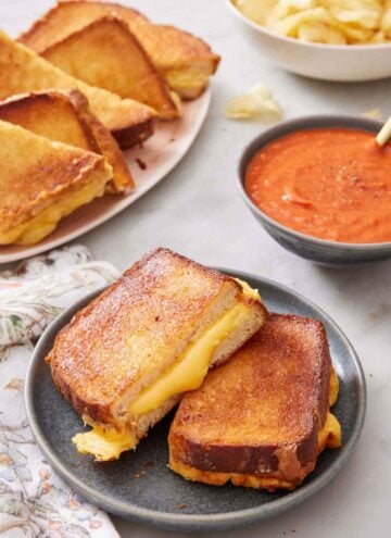A plate with air fryer grilled cheese cut in half with more on a platter in the background along with a bowl of tomato soup.