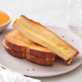 A plate with an air fryer grilled cheese cut in half and slightly stacked on top of each other. A drink and tomato soup in the background.