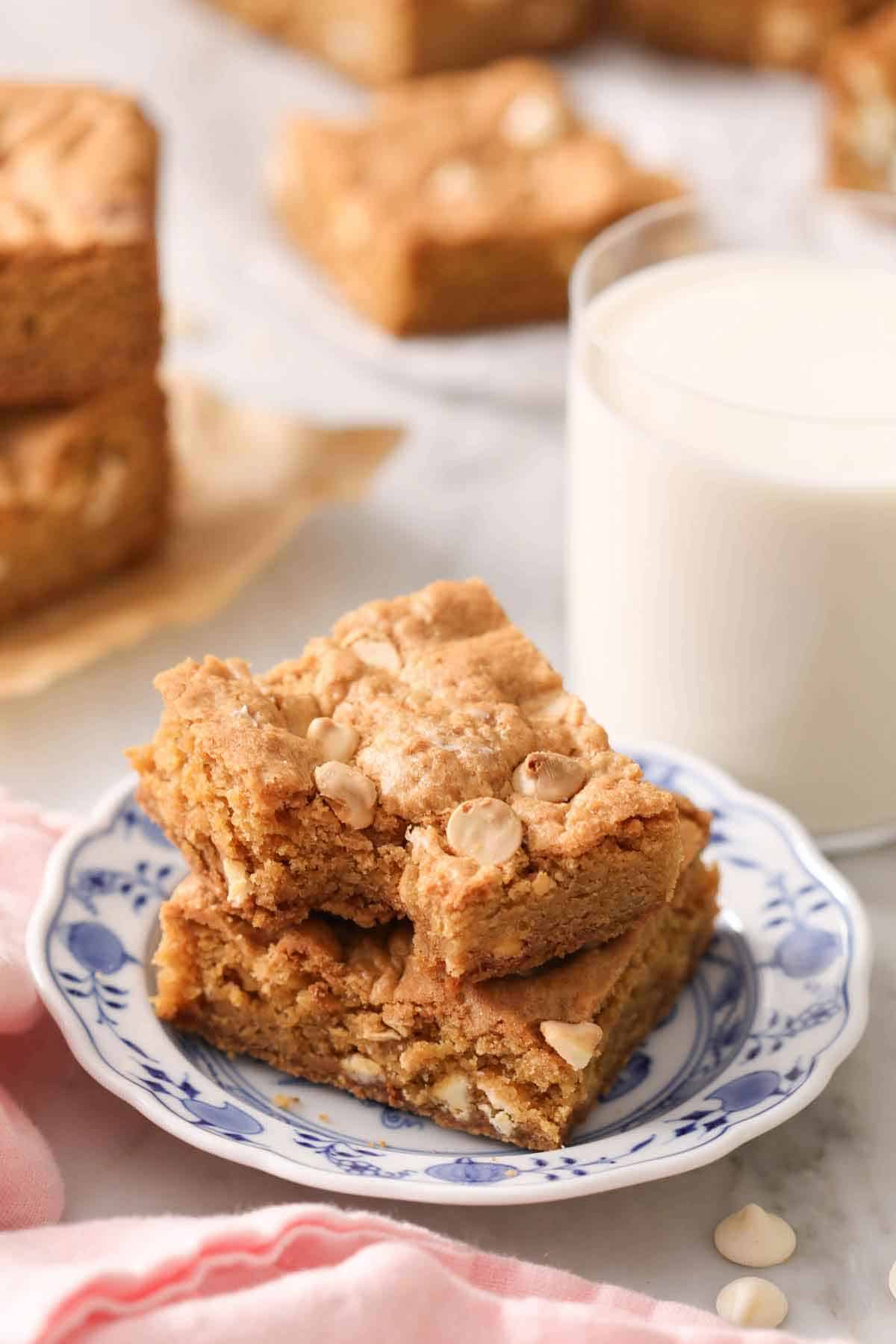 A plate with two blondies, one with a bite taken out, with a glass of milk behind it.