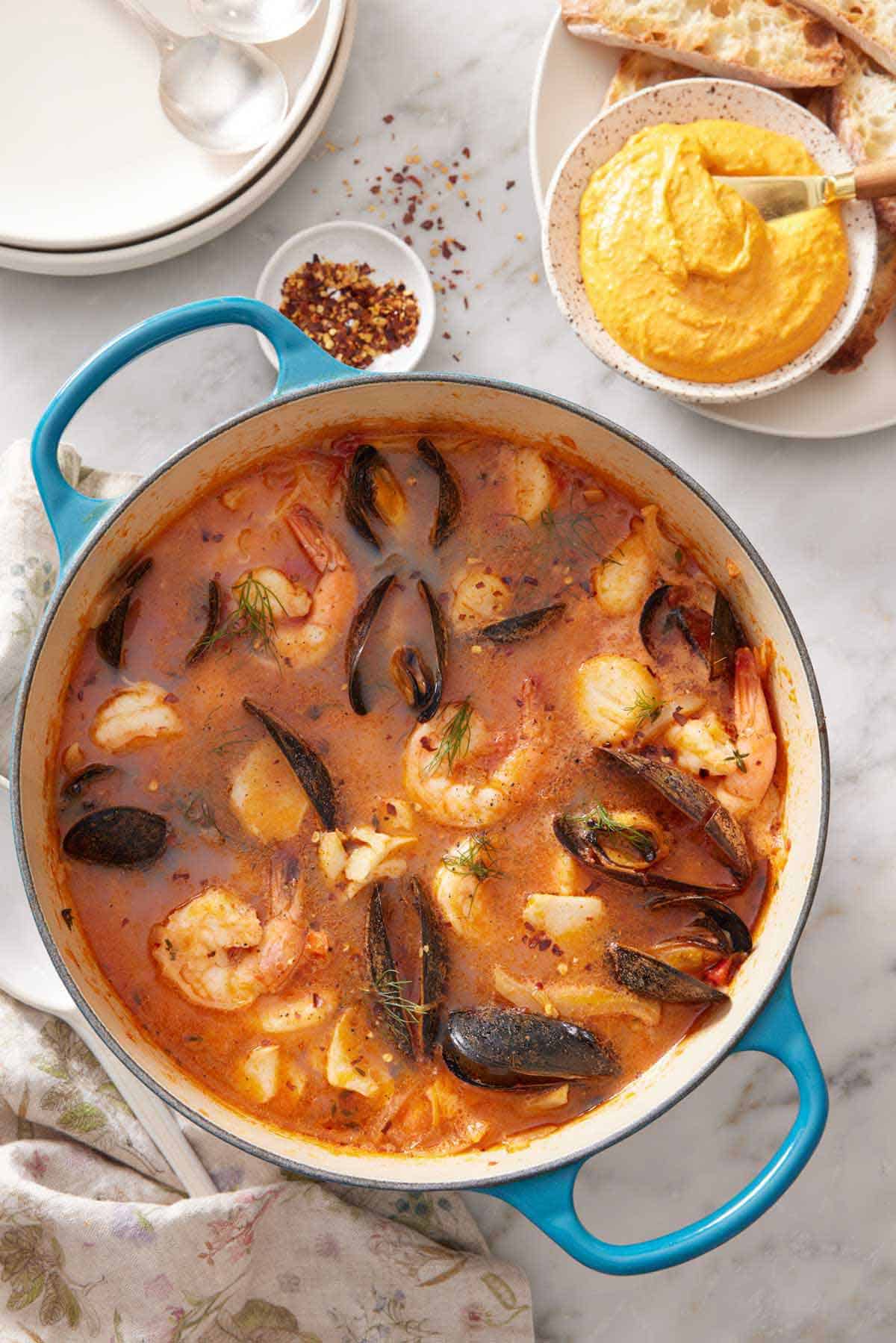 Overhead view of bouillabaisse in a large pot with a bowl of rouille and bread off to the side.