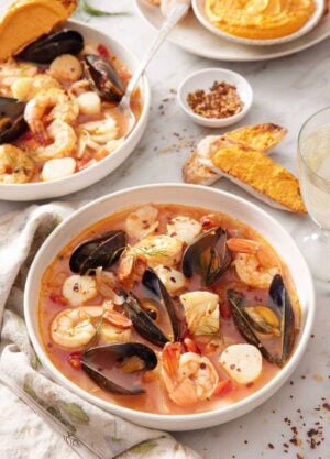 Two bowls of bouillabaisse, one in front, and bread with rouille spread on it in the background along with a bowl of rouille.