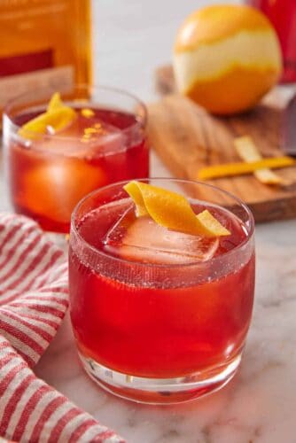 A glass of boulevardier with an orange twist on top. A second glass in the background along with an orange.