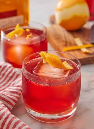 A glass of boulevardier with an orange twist on top. A second glass in the background along with an orange.