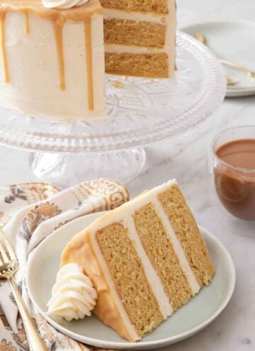 A slice of butterscotch cake on a plate with the rest of the cake in the background on a cake stand.