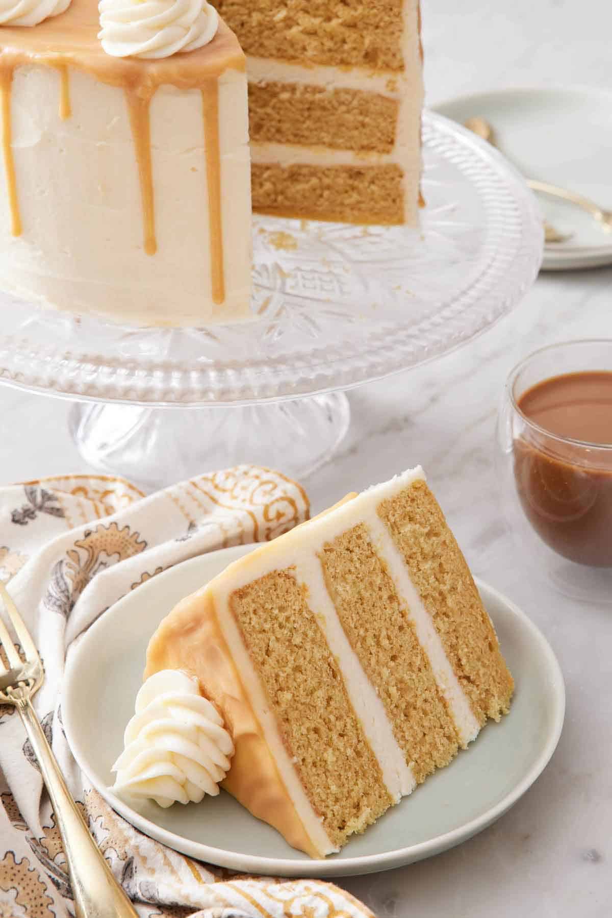 A slice of butterscotch cake on a plate with the rest of the cake in the background on a cake stand.