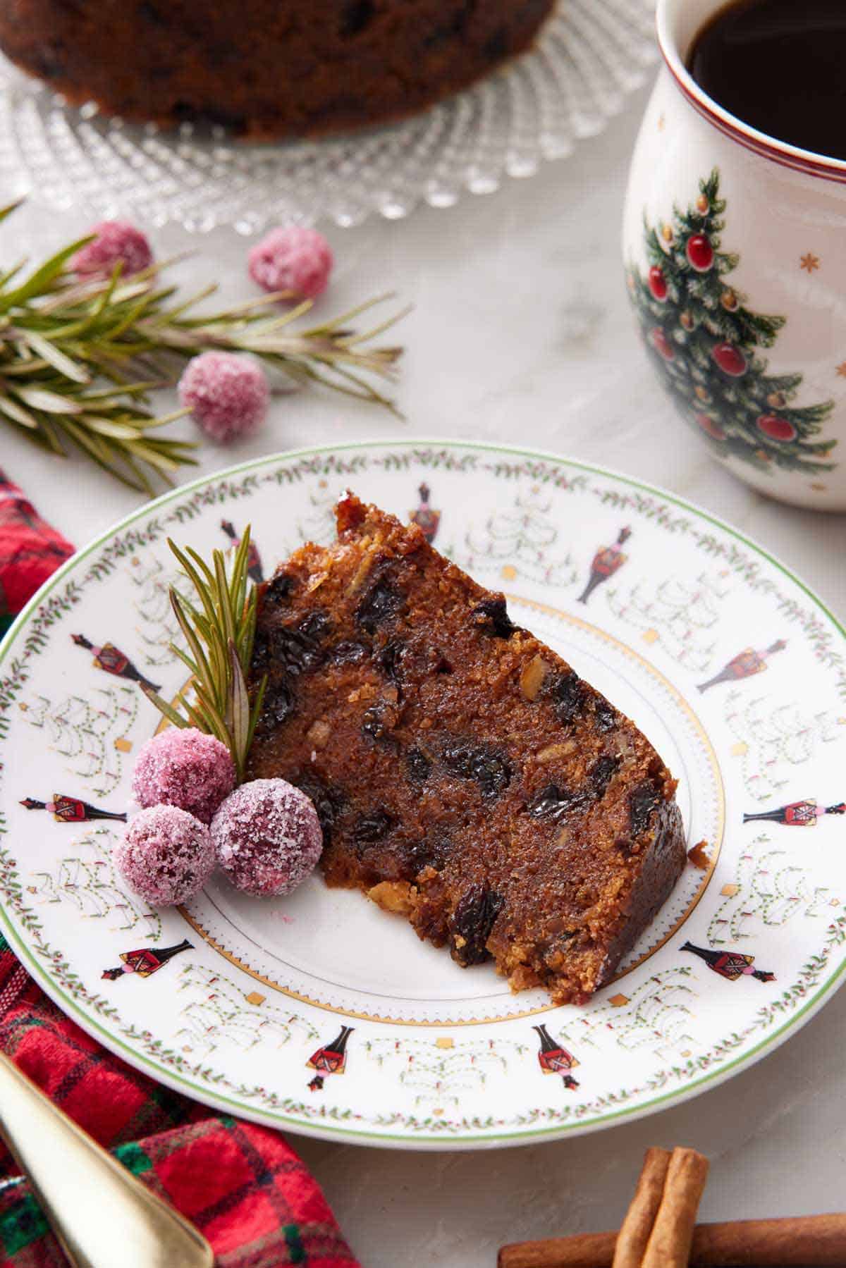A plate with a slice of Christmas pudding with some sugared cranberries and rosemary on the side. A mug of coffee in the background.
