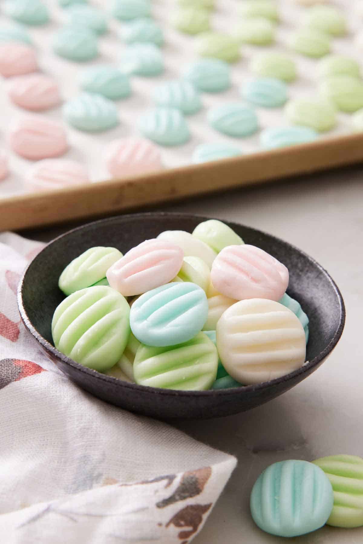 A small bowl of cream cheese mints with a sheet pan with more in the background.