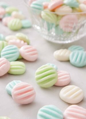 Multiple cream cheese mints in different colors on a marble surface with a bowl with more in the background.
