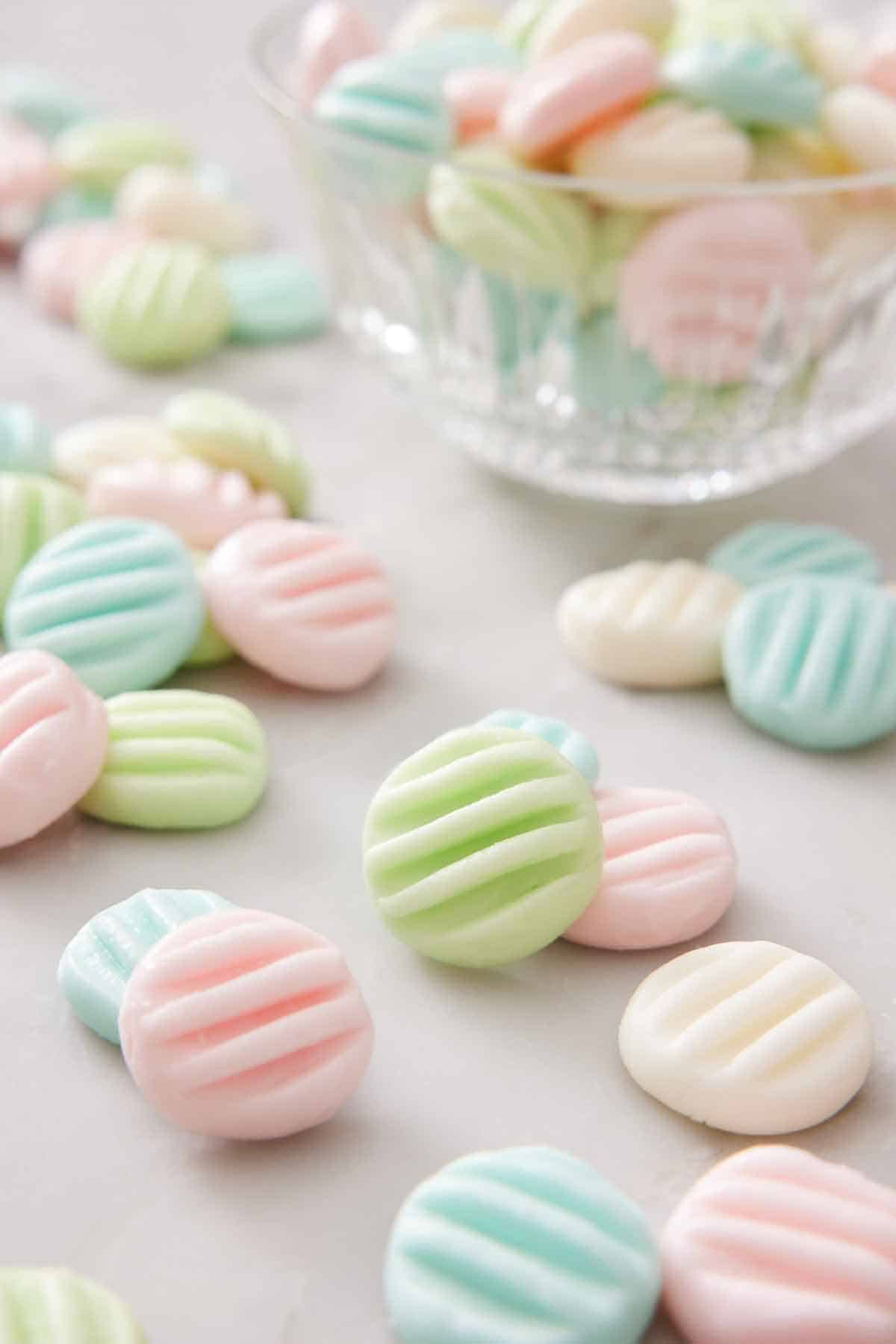 Multiple cream cheese mints in different colors on a marble surface with a bowl with more in the background.