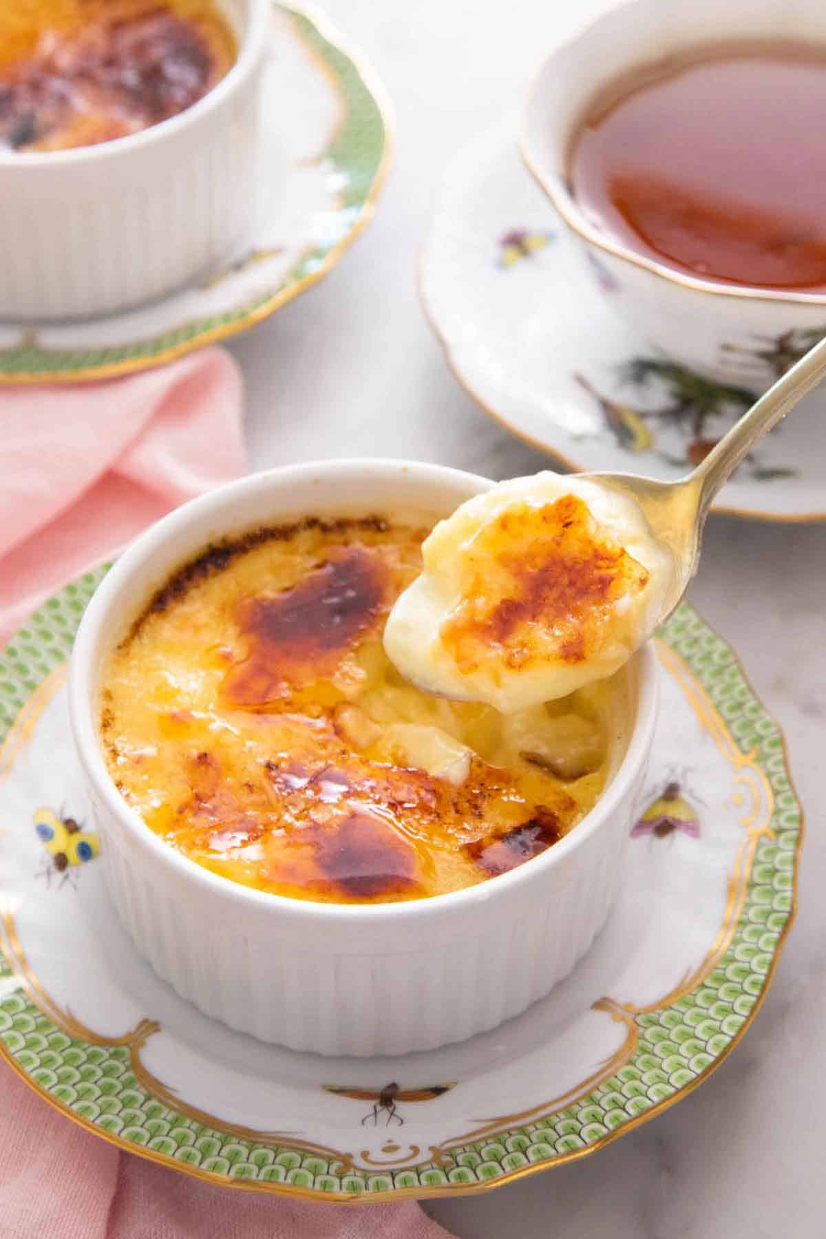 A spoonful of crème brûlée lifted from the ramekin with a cup of tea in the background.