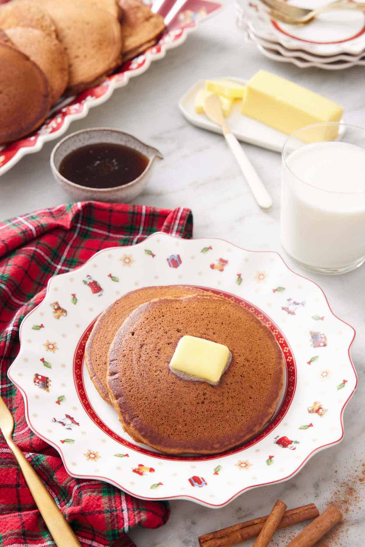 A plate with two gingerbread pancakes with a knob of butter on top. A glass of milk in the back along with butter.