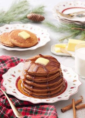 A plate with a stack of gingerbread pancakes with syrup dripping down and butter on top. More pancakes in the background.