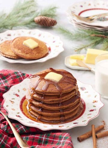 A plate with a stack of gingerbread pancakes with syrup dripping down and butter on top. More pancakes in the background.