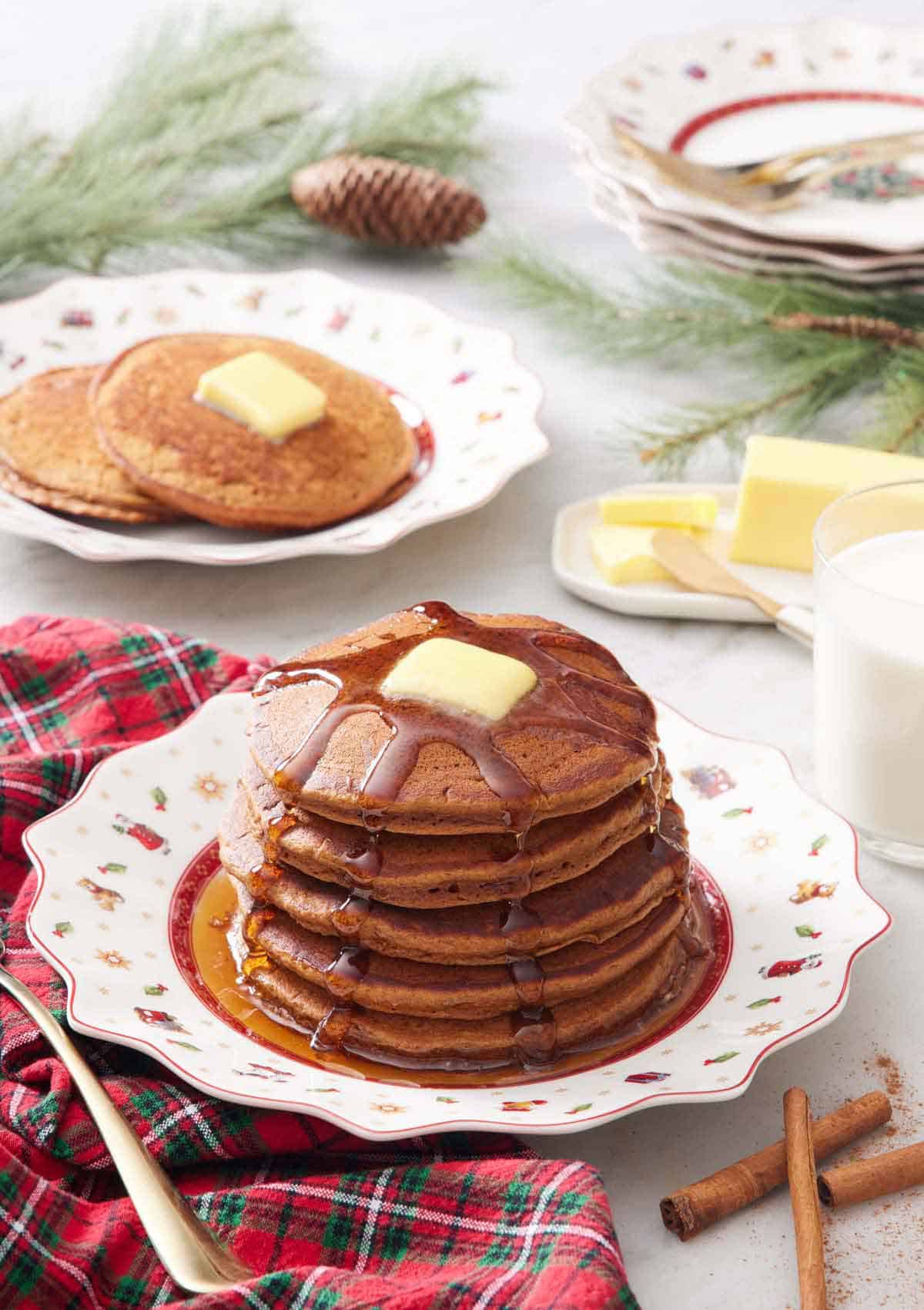 A plate with a stack of gingerbread pancakes with syrup dripping down and butter on top. More pancakes in the background.