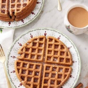 Overhead view of a large gingerbread waffle with a dusting of powdered sugar on top. A stack of waffles in the background along with a coffee and stick of butter.