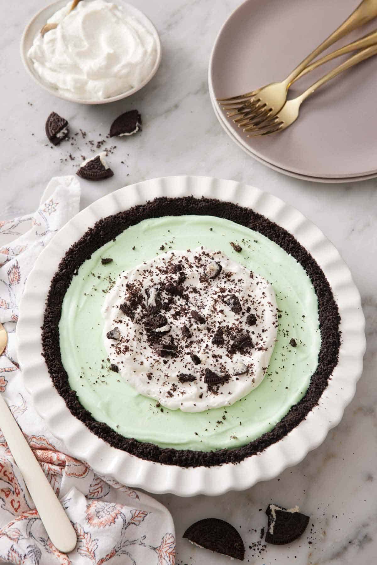 A grasshopper pie in a white baking dish topped with whipped cream and crushed Oreos. A stack of plates and forks in the background along with oreos and whipped cream.