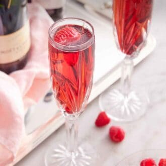 Pinterest graphic of two champagne flutes of Kir Royale with a bowl of raspberries and the bottles of alcohol in the back.