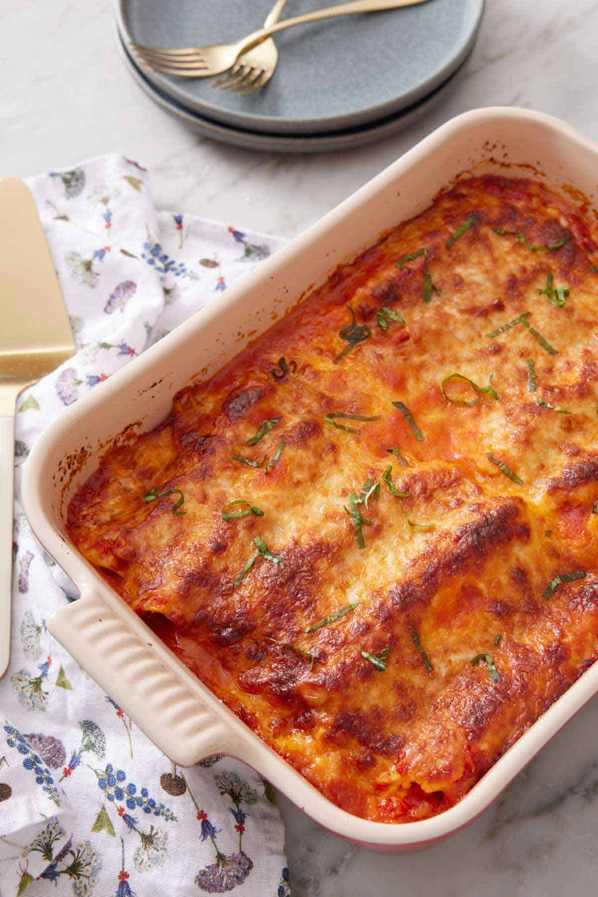 A baking dish of manicotti with a stack of plates and forks in the background.